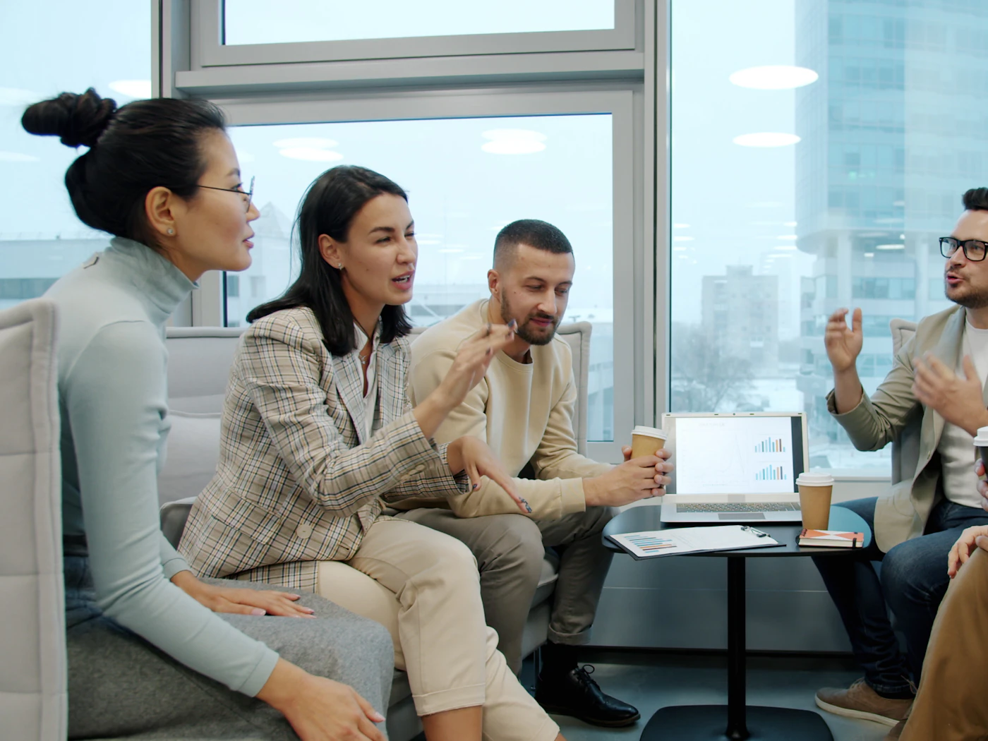 A diverse team in an animated discussion on lounge seating by a sunny office window, coffee in hand