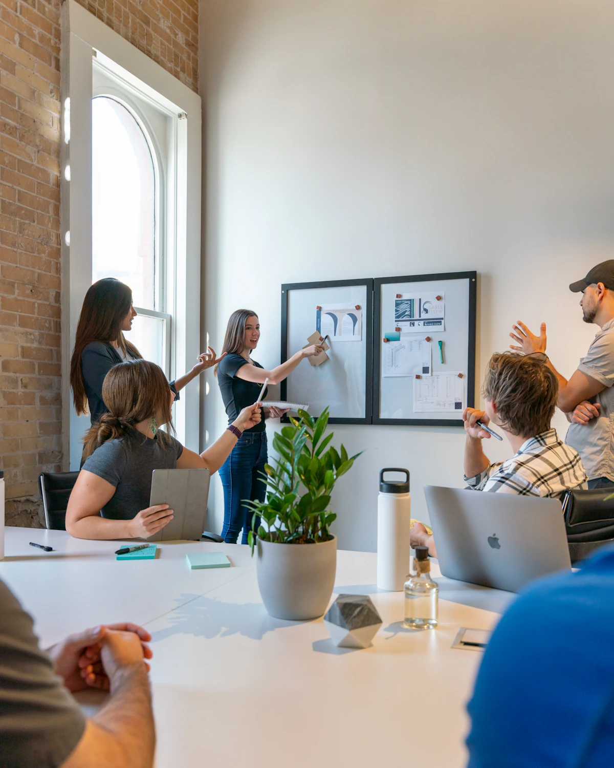 A team gathered around a white table in a bright brick-walled room while a colleague presents plans at a wall board